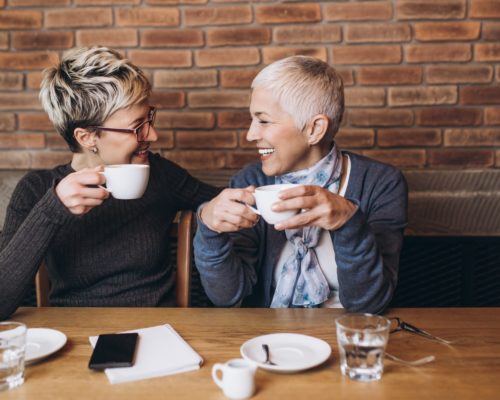 Two women smiling at each other sitting at table holding cups of coffee.