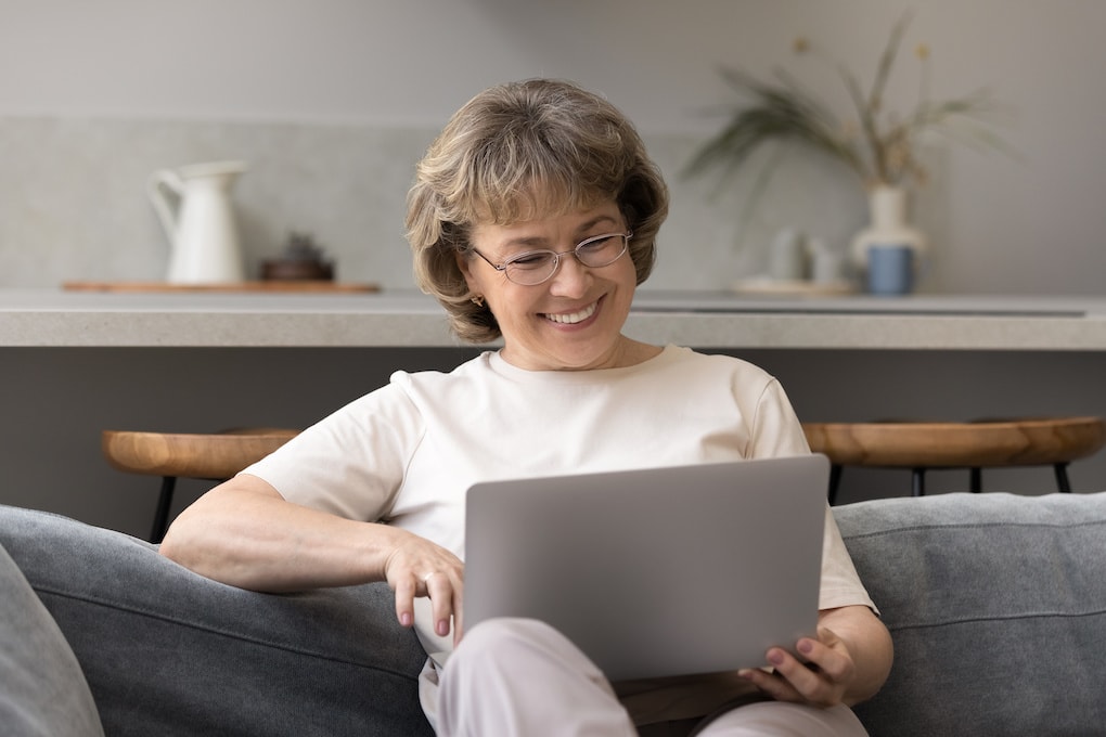 Elderly woman smiling on couch learning how to schedule a telehealth appointment form her laptop